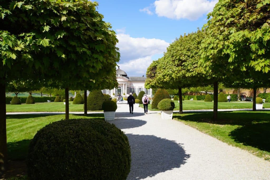 Ein gepflegter Kiesweg führt durch den Garten von Stift Melk, gesäumt von akkurat geschnittenen Kugelbäumen und Heckenfiguren, während im Hintergrund das barocke Gartenpavillon unter blauem Himmel zu sehen ist.