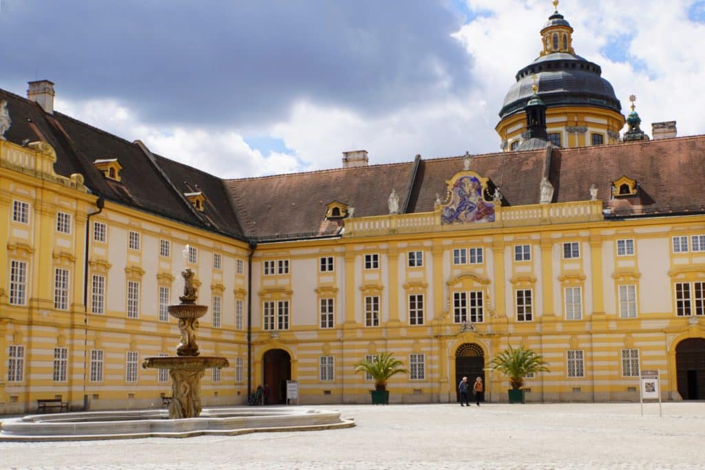Der sonnige Innenhof von Stift Melk zeigt die gelb-weiße Barockfassade mit einem zentralen Brunnen im Vordergrund und der markanten Stiftskuppel im Hintergrund.