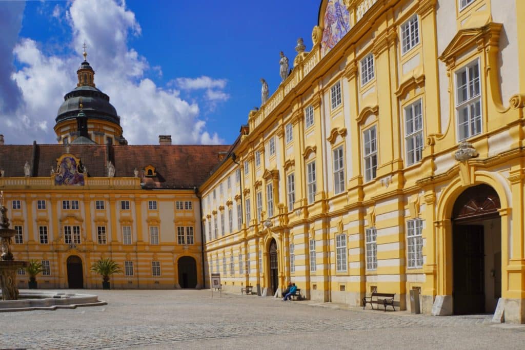 Der sonnendurchflutete Innenhof des Stift Melk zeigt die prachtvolle gelbe Barockfassade mit zahlreichen Fenstern und Statuen unter einem blauen Himmel mit weißen Wolken. Im Hintergrund ragt die markante dunkle Kuppel der Stiftskirche über die rötlichen Ziegeldächer hinaus.