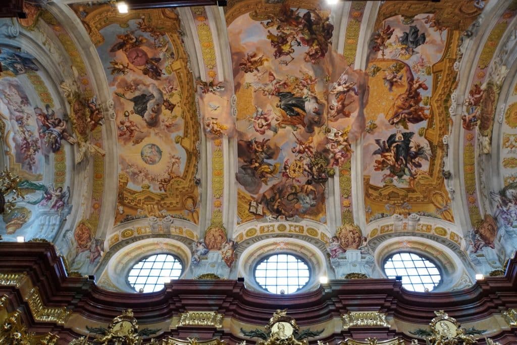 Prachtvolle, barocke Deckenfresken in der Stift Melk Kirche zeigen zahlreiche religiöse Figuren und Engel in einem wolkenverhangenen Himmel, eingerahmt von goldfarbenen Stuckaturen und runden Fenstern.