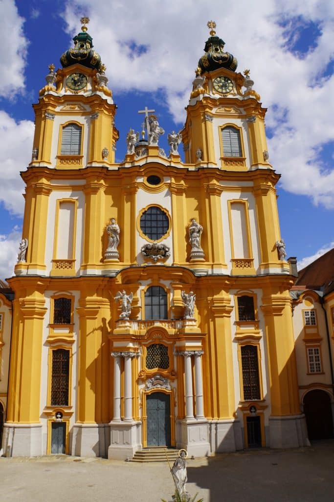 Die prachtvolle gelb-weiße Barockfassade der Stiftskirche Melk mit ihren zwei markanten Zwiebeltürmen, zahlreichen Heiligenstatuen und einem zentralen Portal unter einem strahlend blauen Himmel.