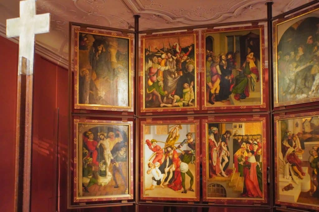 Ein mehrteiliges, spätgotisches Altarbild im Museum des Stifts Melk zeigt in detaillierten Tafelgemälden verschiedene Szenen aus der Passionsgeschichte Christi, flankiert von einem schlichten Holzkreuz im Vordergrund.