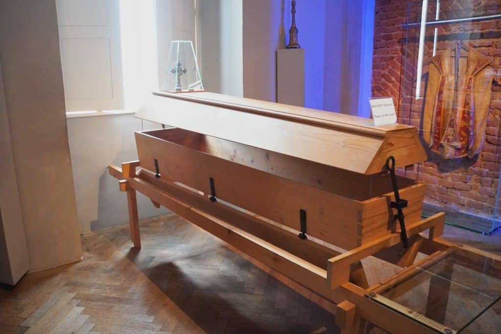Ein hölzerner Klappsarg aus dem Stift Melk steht auf einem Holzgestell in einem Ausstellungsraum, wobei der Boden des Sarges nach unten geöffnet ist. Im Hintergrund sind eine gemauerte Ziegelwand, ein religiöses Gewand in einer Vitrine und ein Fenster zu sehen.