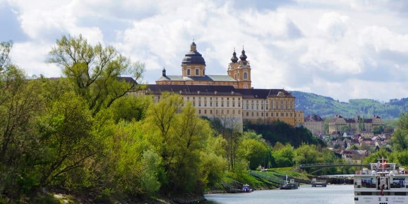 Blick vom Wasser der Donau auf das gelbe Barockgebäude des Stift Melk, das auf einem Felsen über grünen Bäumen thront, während im Vordergrund ein Flusskreuzfahrtschiff fährt.