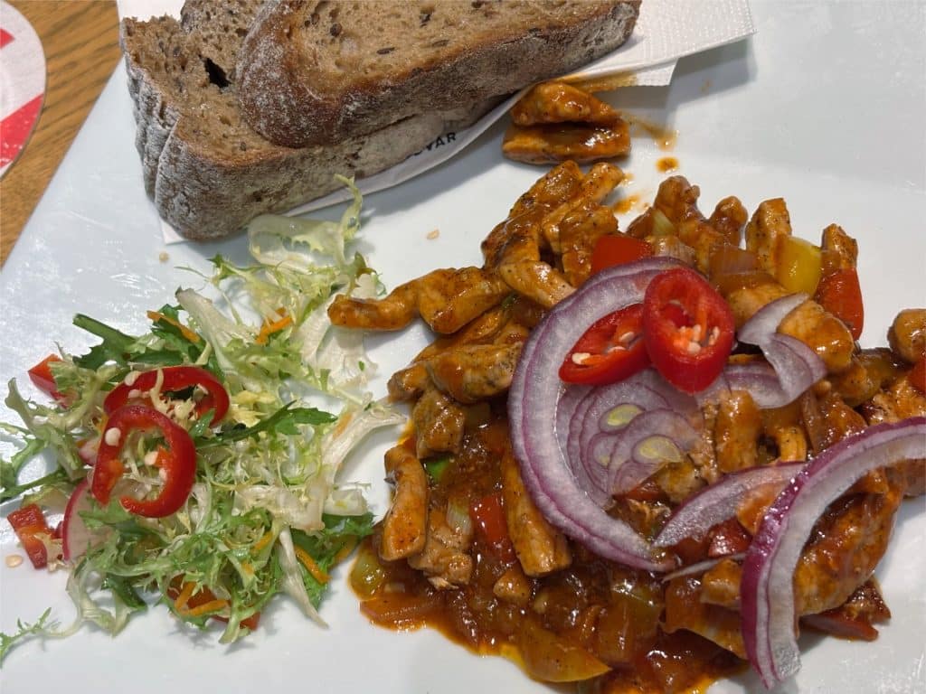 Ein Teller mit einem herzhaften Gericht aus der Gastronomie der Budweiser Brauerei, bestehend aus würzigem Fleischgeschnetzeltem in Sauce, garniert mit roten Zwiebelringen und Peperoni, serviert mit zwei Scheiben dunklem Brot und einem frischen Beilagensalat.