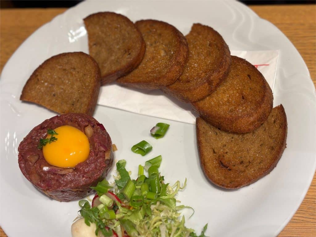 Ein Teller mit Rinder-Tatar, garniert mit einem Eigelb und frischen Kräutern, serviert mit gerösteten dunklen Brotscheiben und einem kleinen Beilagensalat in einem Restaurant in Budweis.