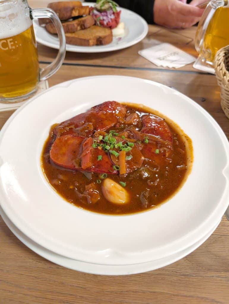 Ein Teller mit in dunkler Soße eingelegten Gulasch, garniert mit Schnittlauch und Knoblauch, serviert im Restaurant Solnice in Budweis neben einem Glas Bier.