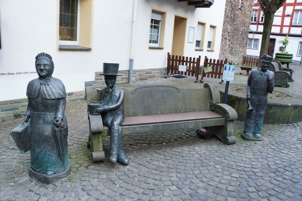 Ein Kunstwerk in Cochem zeigt drei lebensgroße Bronzestatuen historisch gekleideter Personen, die um eine steinerne Sitzbank auf einem gepflasterten Platz angeordnet sind.