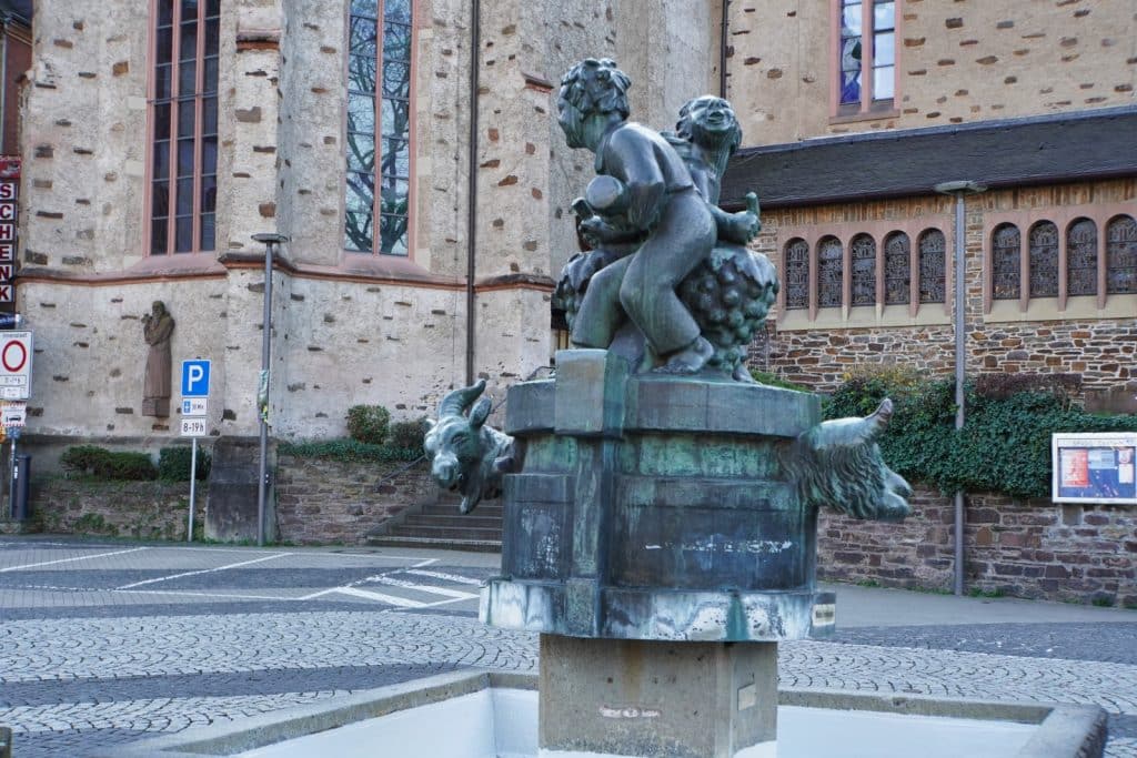 Eine bronzene Skulptur des brunnens in Cochem zeigt zwei spielende Kinder auf einem Weinfass, das mit Tierköpfen wie einer Ziege verziert ist, vor der Kulisse einer historischen Kirche.