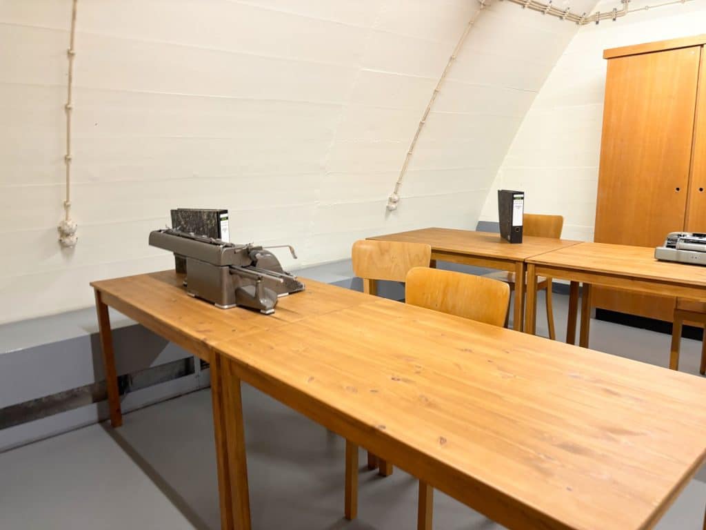 Ein schlicht eingerichteter Arbeitsraum im Bundesbankbunker Cochem mit mehreren Holztischen, einfachen Stühlen, Aktenordnern und einer historischen Schreibmaschine unter einer gewölbten Betondecke.