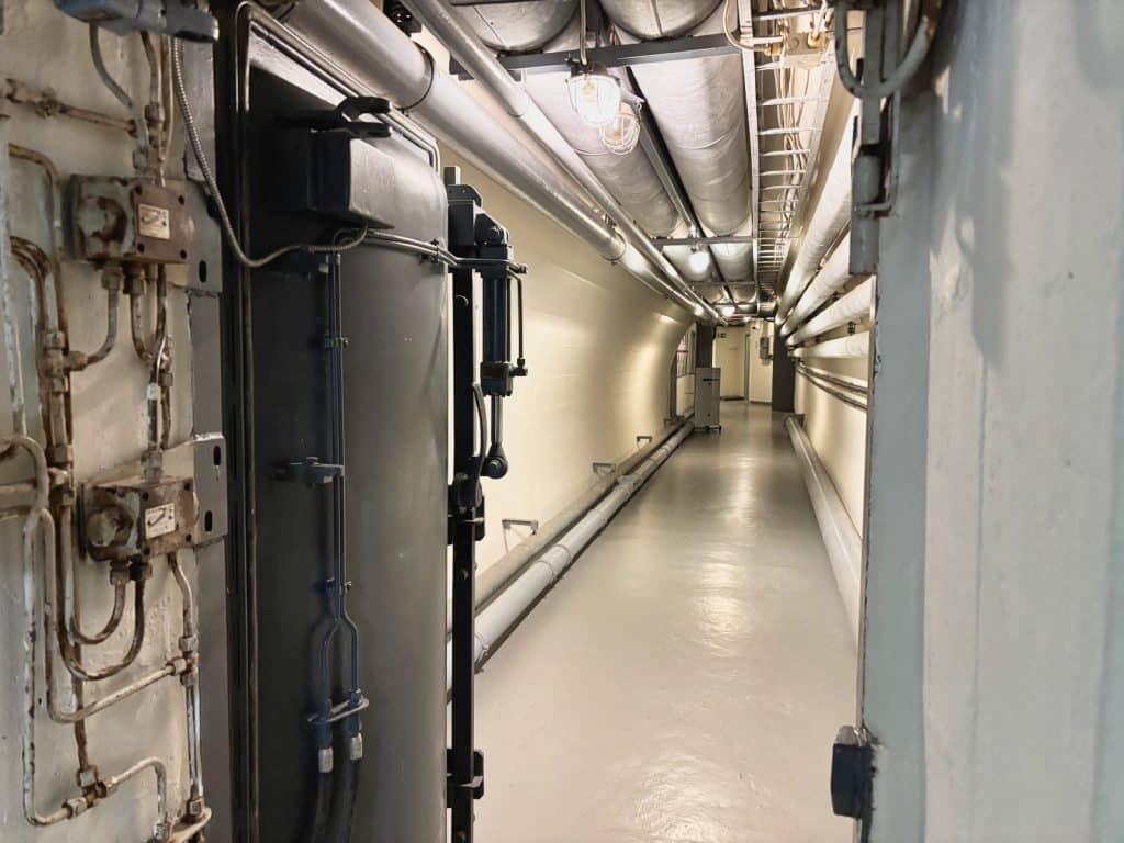 Ein langer, schmaler Gang im ehemaligen Bundesbank-Bunker in Cochem mit einer schweren Panzertür im Vordergrund und zahlreichen Rohrleitungen an der Decke und den Wänden.