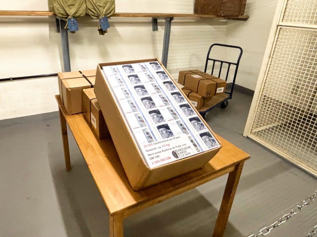 Ein offener Pappkarton voller Geldschein-Bündel steht auf einem Holztisch im Cochem Bunker, daneben weitere geschlossene Kisten und ein Transportwagen im Hintergrund.