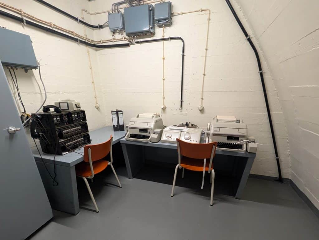 Ein schlicht eingerichteter Raum der Kommunikationszentrale im Bundesbankbunker Cochem mit zwei grauen Arbeitstischen, auf denen historische Fernschreiber, ein Telefon-Vermittlungspult und zwei orangefarbene Stühle stehen. An den weißen Wänden verlaufen zahlreiche elektrische Leitungen und graue Schaltkästen unter einer gewölbten Decke.