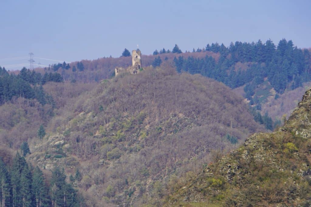 Die Ruine der Winneburg bei Cochem thront auf einem bewaldeten Hügel vor einer Kulisse aus dichten Nadelwäldern unter einem klaren, hellblauen Himmel.