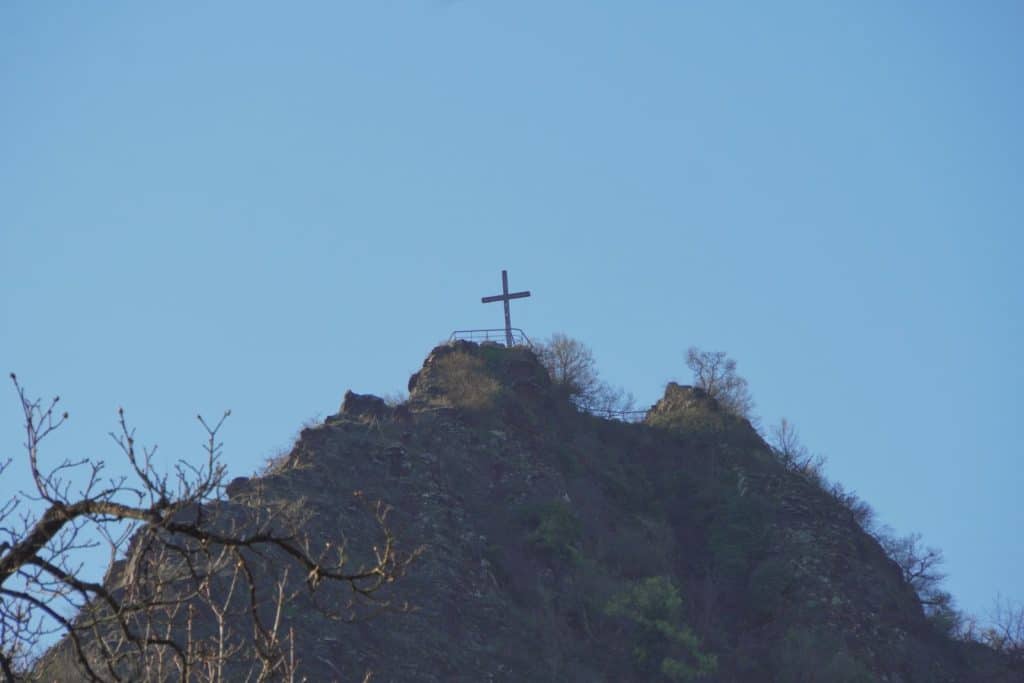 Ein hölzernes Gipfelkreuz steht auf der felsigen Spitze einer Anhöhe über Cochem vor einem klaren, blauen Himmel. Im Vordergrund ragen kahle Äste in das Bild, während der steile, dunkle Felshang vereinzelt mit grünem Gebüsch bewachsen ist.