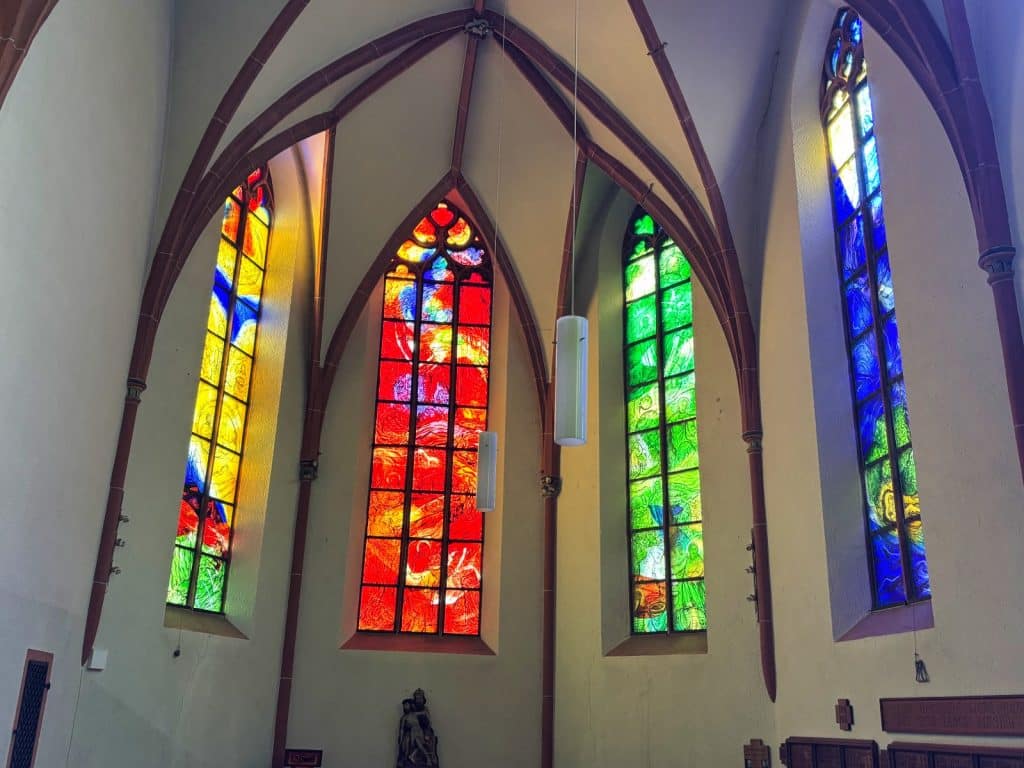 Vier hohe, spitzbogige Buntglasfenster in den Farben Gelb, Rot, Grün und Blau tauchen den Altarraum einer Kirche in Cochem in farbiges Licht. Die Fenster sind in eine helle Wand mit gotischen Gewölberippen eingelassen, während davor schlichte, moderne Zylinderleuchten von der Decke hängen.