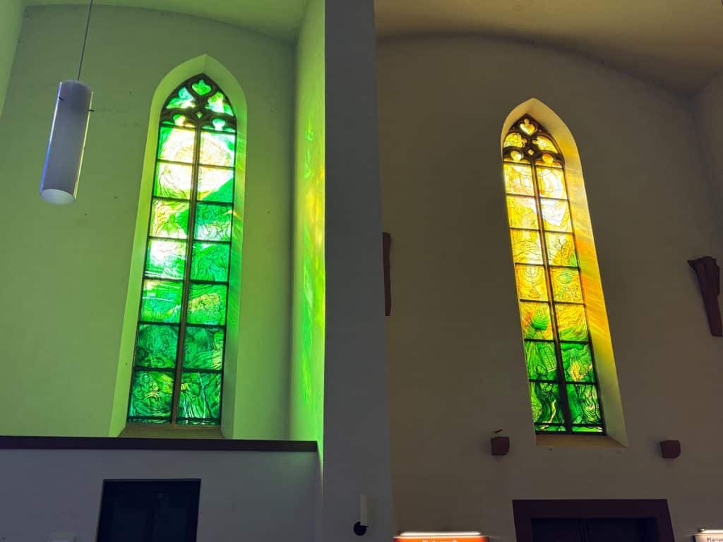 Zwei hohe, spitzbogige Buntglasfenster in der Kirche von Cochem zeigen religiöse Motive in leuchtenden Grün- und Gelbtönen, deren farbiges Licht auf die umliegenden weißen Innenwände fällt.