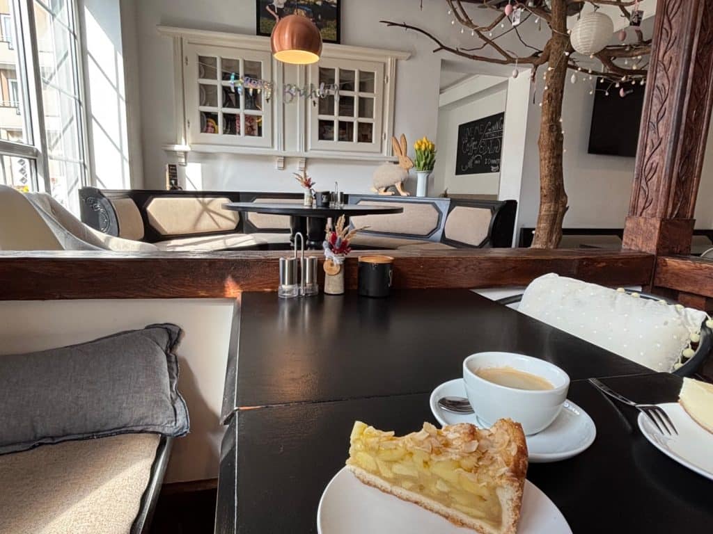 Ein Stück Apfelkuchen und eine Tasse Kaffee stehen auf einem dunklen Tisch in einem gemütlich eingerichteten Café in Cochem, das mit rustikalen Holzmöbeln und österlicher Dekoration gestaltet ist.