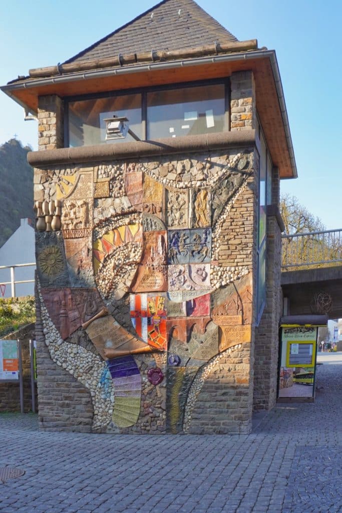 Ein zweistöckiger Steinturm an der Brücke in Cochem, dessen Fassade mit einem großflächigen, farbenfrohen Mosaik-Kunstwerk aus Reliefs, Wappen und historischen Motiven verziert ist.