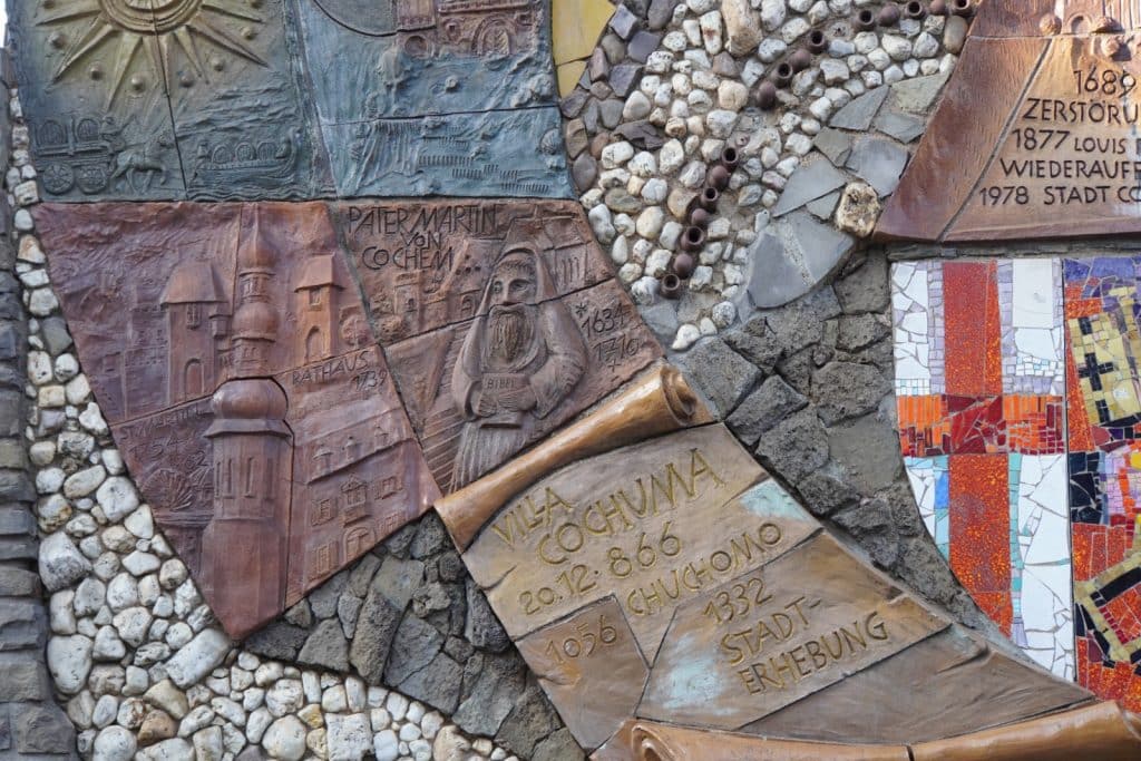 Detailansicht eines Mosaik-Kunstwerks in Cochem, das historische Szenen, Persönlichkeiten wie Pater Martin von Cochem und wichtige Jahreszahlen der Stadtgeschichte in reliefartigen Keramik- und Steinelementen darstellt.