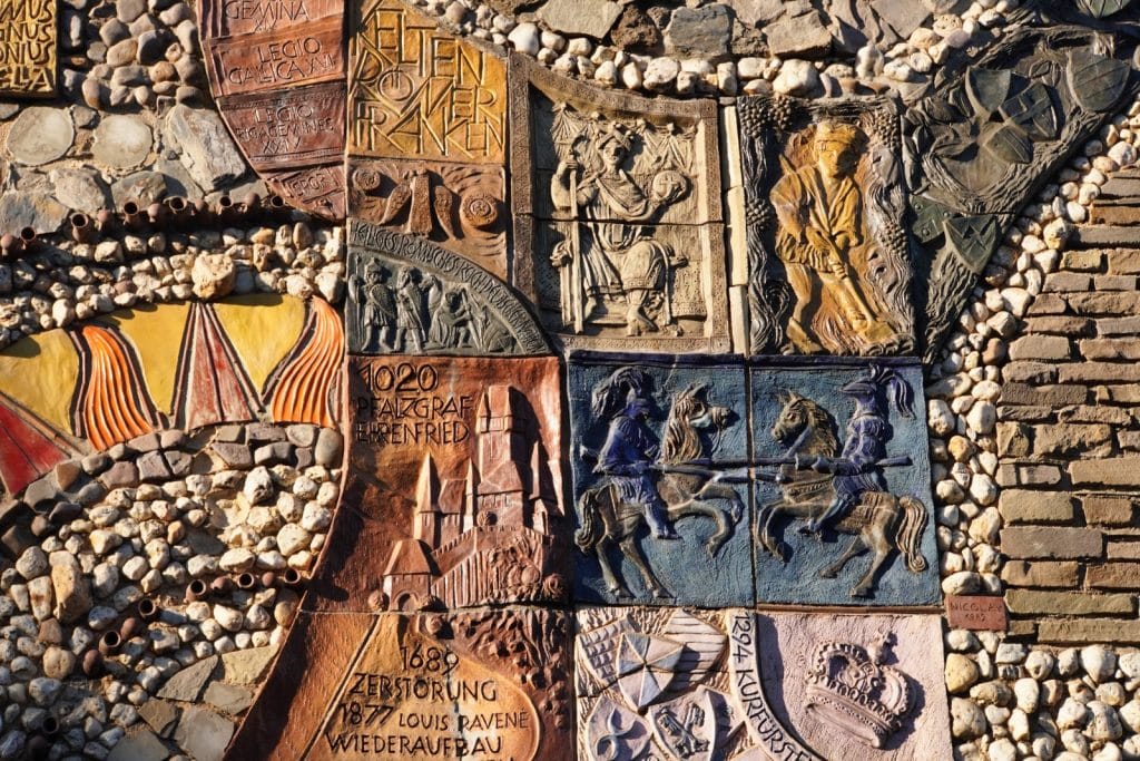 Ein detailreiches Mosaik-Kunstwerk in Cochem zeigt verschiedene historische Szenen und Inschriften in Steinreliefs, darunter Ritterturniere, Burgen und Wappen, die die Geschichte der Region illustrieren.