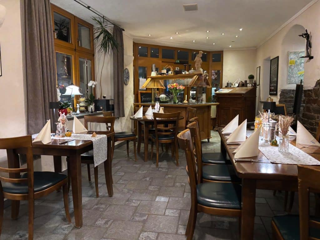 Der gemütliche Gastraum des Restaurants Onkel Willi in Cochem ist mit rustikalen Holztischen, gepolsterten Stühlen und hellen Servietten eingedeckt und wird durch warmes Licht sowie große Fensterfronten beleuchtet.