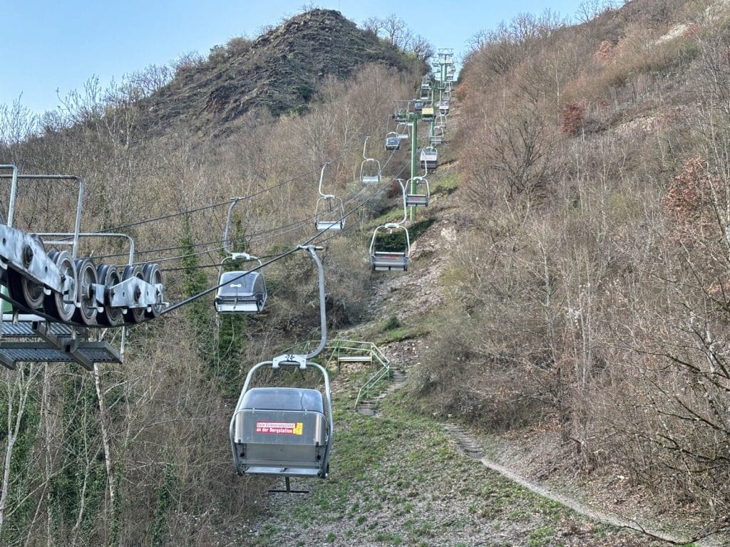 Ein Sessellift in Cochem führt mit zahlreichen silbernen Gondeln einen steilen, bewaldeten Hang hinauf zu einer Bergstation.