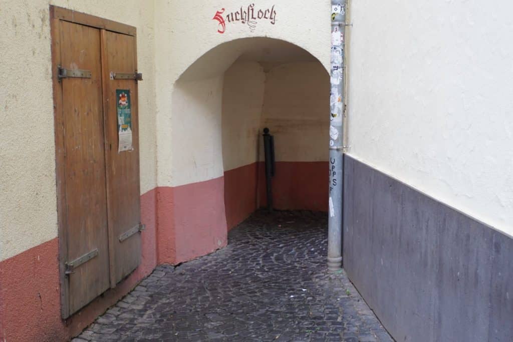 Ein schmaler, gepflasterter Durchgang führt unter einem Rundbogen hindurch, über dem in roter Frakturschrift der Name „Fuchsloch“ steht, ein historisches Stadttor in Cochem. Links neben dem Eingang befindet sich eine rustikale Holztür mit Metallbeschlägen an einer hellgelben Hauswand.