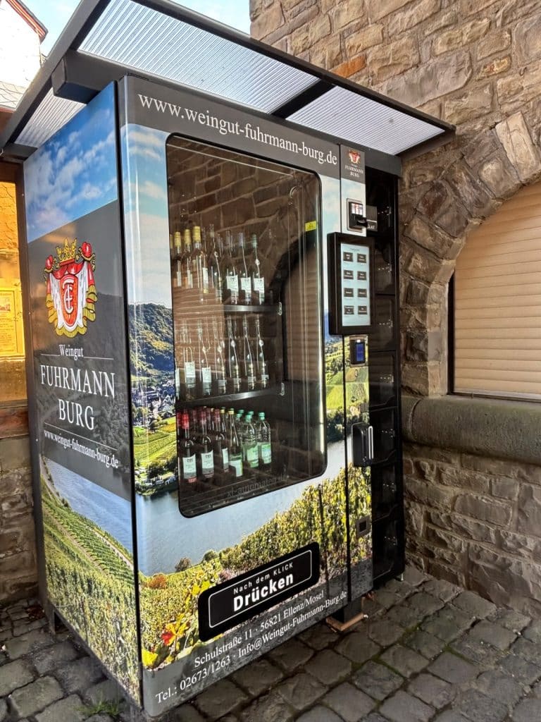 Ein moderner Weinautomat des Weinguts Fuhrmann-Burg steht vor einer rustikalen Steinwand in Cochem und bietet verschiedene Flaschenweine zur Selbstbedienung an. Das Gerät ist mit großflächigen Weinberg-Motiven und dem Logo des Weinguts gestaltet.