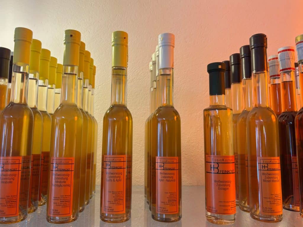 Mehrere Reihen schlanker Glasflaschen mit verschiedenen Sorten von der Manufaktur Essigkultur, die mit orangefarbenen Etiketten und unterschiedlich farbigen Verschlusskappen in einem Regal präsentiert werden.