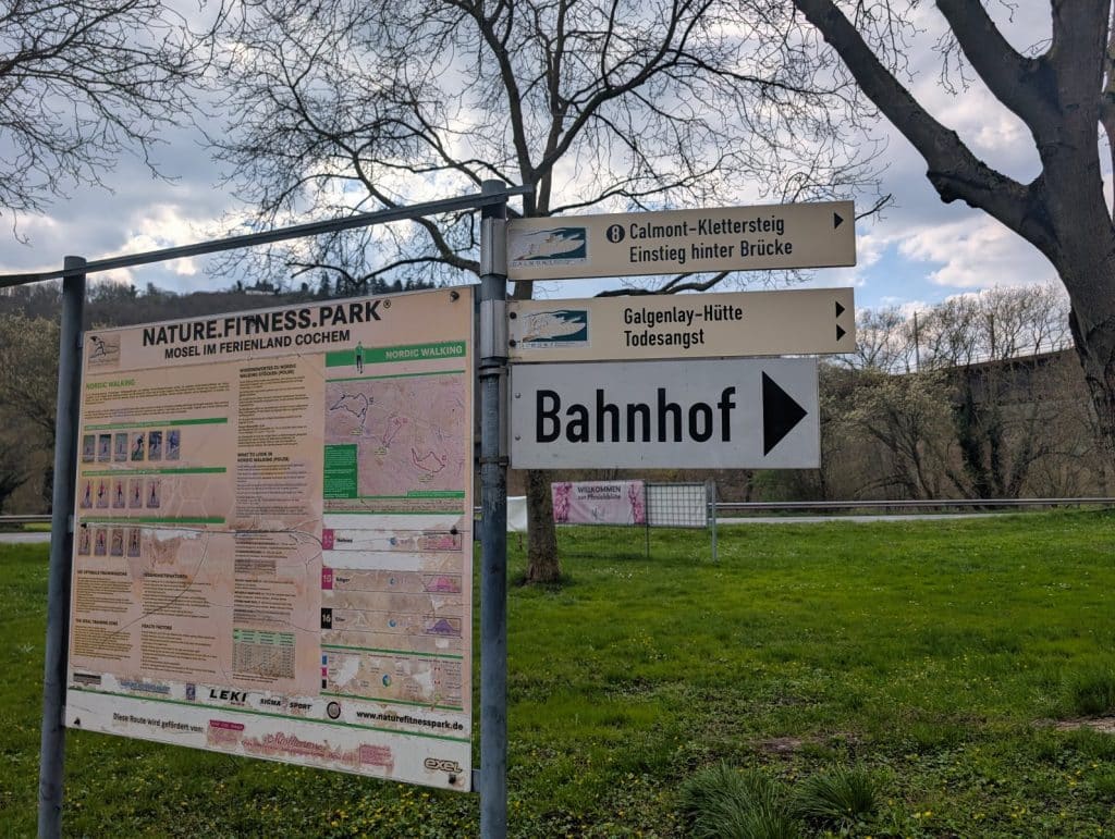 Ein Wegweiser in Eller zeigt mit einem schwarzen Pfeil nach rechts in Richtung „Bahnhof“, während darüber weitere Schilder zum Calmont-Klettersteig und zur Galgenlay-Hütte weisen, flankiert von einer Informationstafel des „Nature.Fitness.Park“.