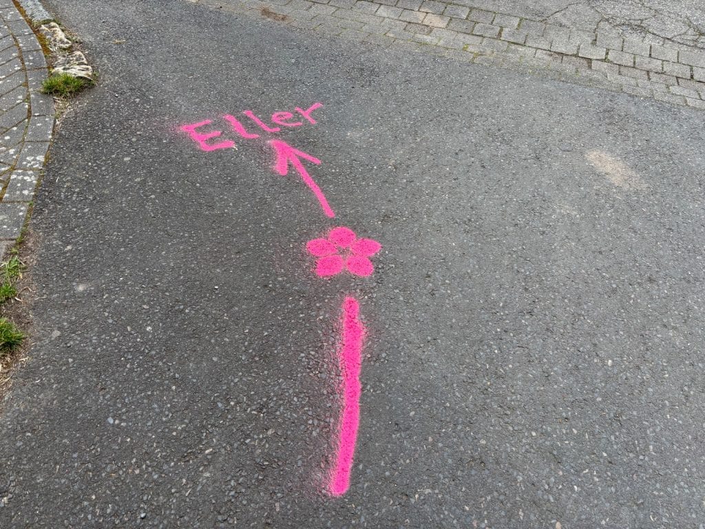 Auf dem Asphalt einer Straße ist eine pinkfarbene Wegmarkierung nach Eller aufgesprüht, bestehend aus einer Linie, einer kleinen Blume und einem Pfeil, der auf den Schriftzug „Eller“ weist.