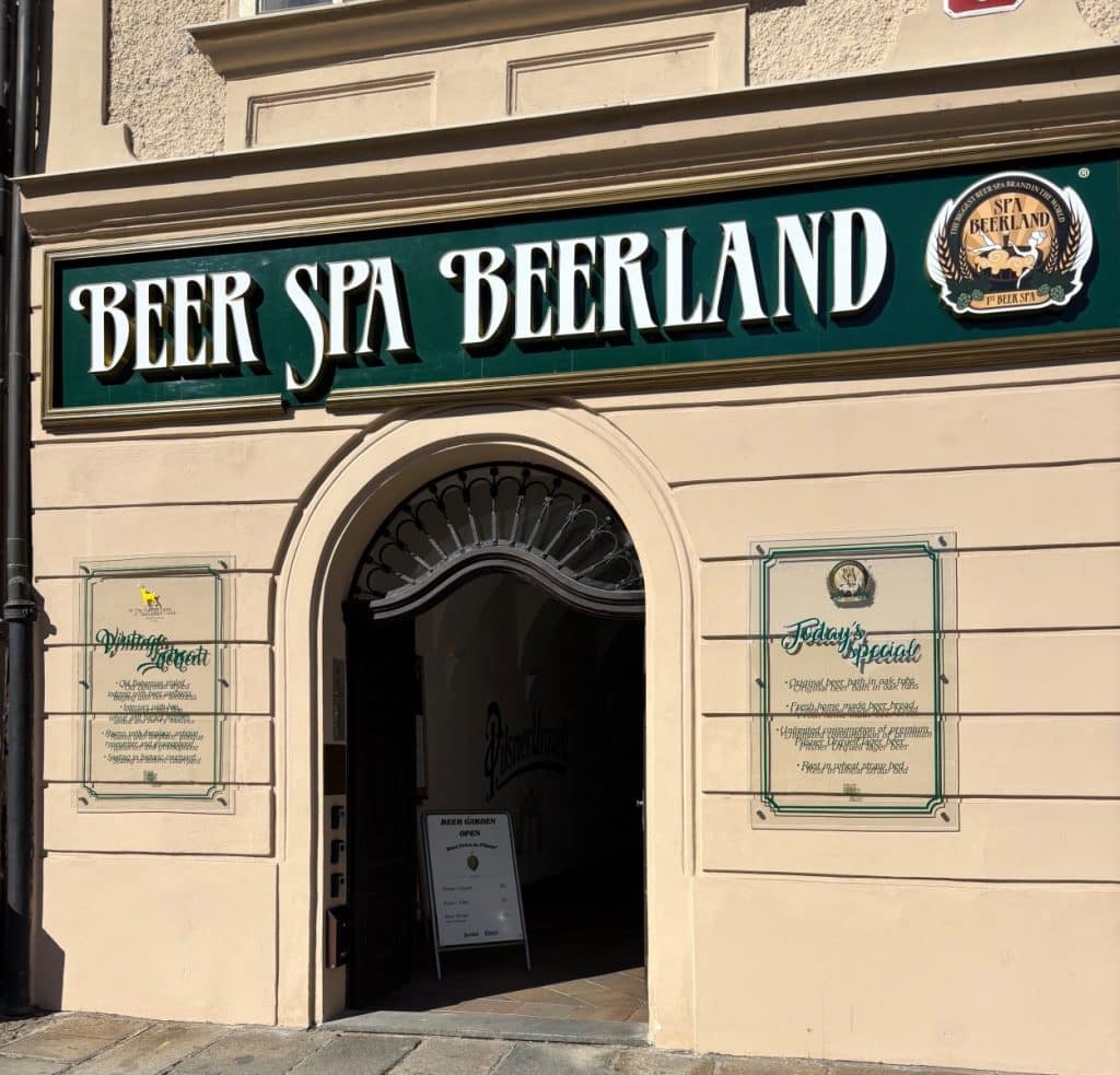 Die Außenansicht des Pilsen Bier Spa Beerland zeigt einen bogenförmigen Eingangsbereich unter einem großen grünen Schild mit weißer Aufschrift und zwei seitlich angebrachten Informationstafeln an der hellen Fassade.