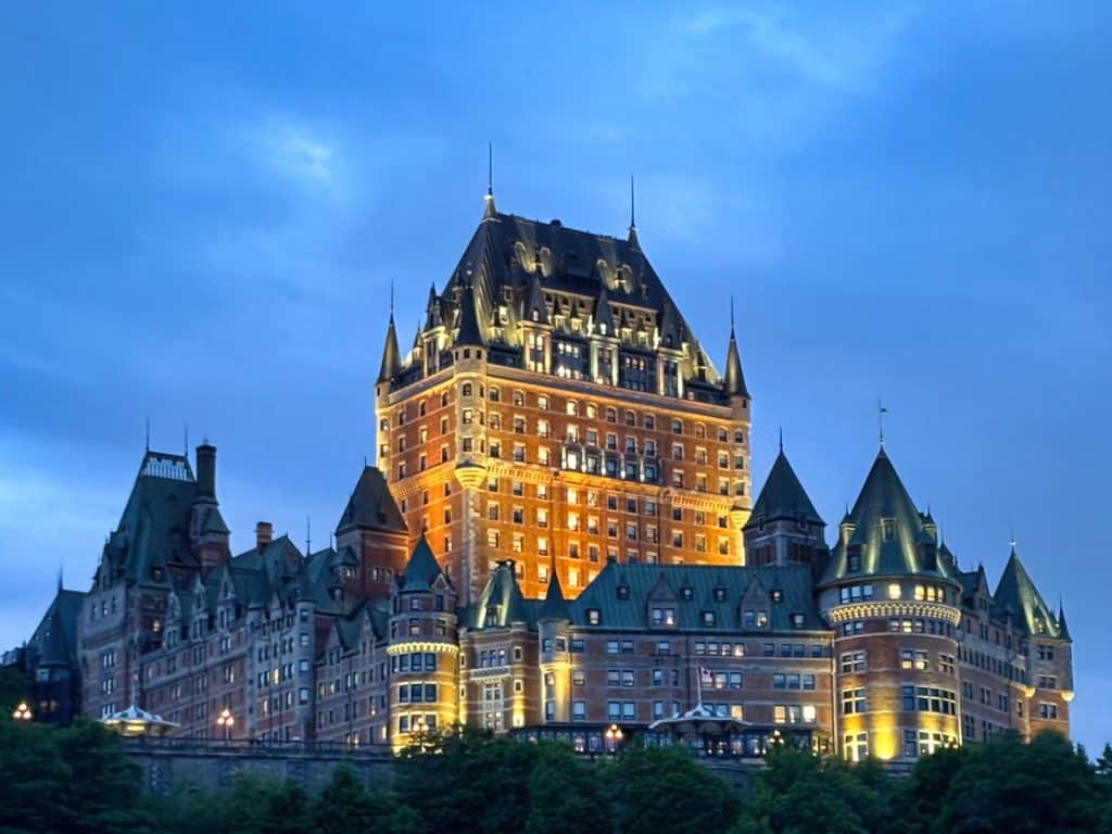 Das beleuchtete Fairmont Le Château Frontenac in Quebec City thront bei blauer Stunde majestätisch über einer grünen Baumreihe vor einem tiefblauen Abendhimmel.