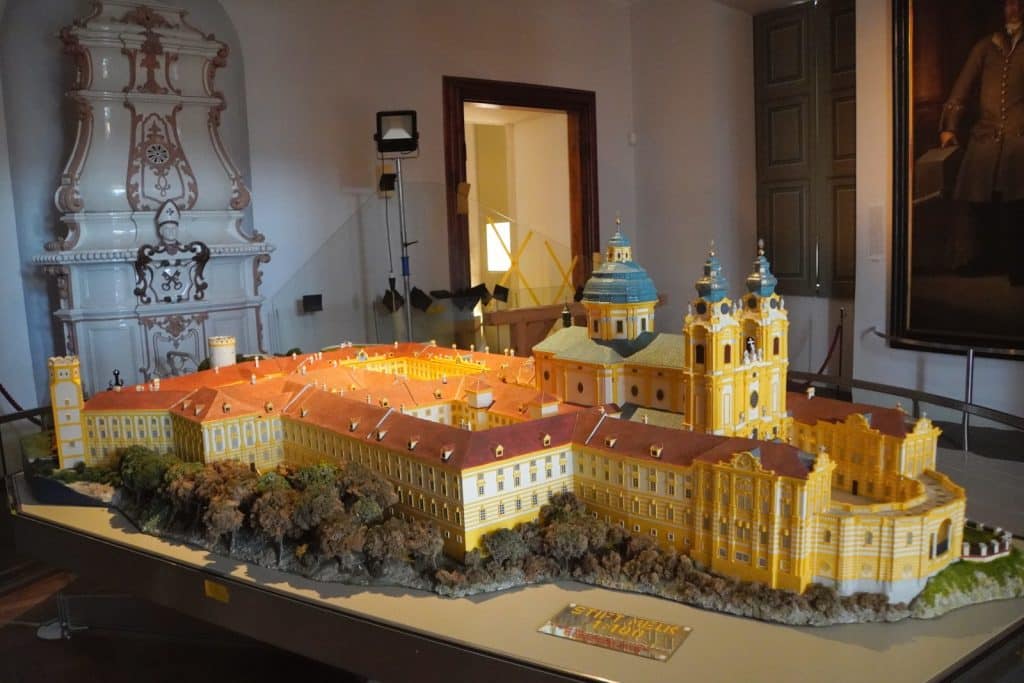 Ein detailliertes Architekturmodell des Stiftes Melk im Maßstab 1:100 steht auf einem Podest in einem Museumsraum mit barocker Einrichtung. Das gelb-weiße Modell zeigt die weitläufige Klosteranlage mit ihrer markanten Kuppelkirche und den Türmen, umgeben von Miniatur-Bäumen auf einem künstlichen Felsen.