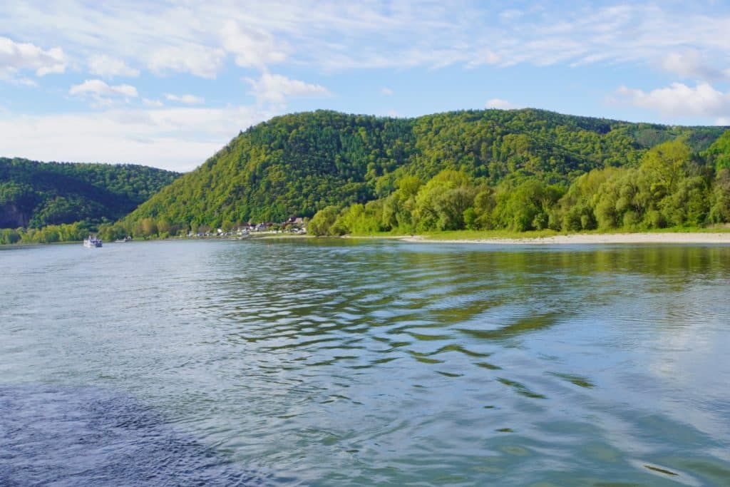 Urlaub in der Wachau: Blick vom Wasser auf die bewaldeten Hügel der Wachau, an deren Ufer ein kleines Dorf und ein helles Flussufer unter leicht bewölktem Himmel liegen.