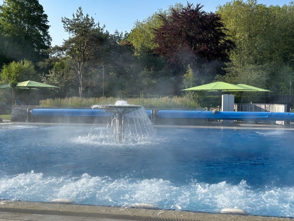 Dampfendes Außenbecken der Therme in Bad Sassendorf mit einem zentralen Wasserpilz, sprudelndem Wasser im Vordergrund und grünen Bäumen im Hintergrund.