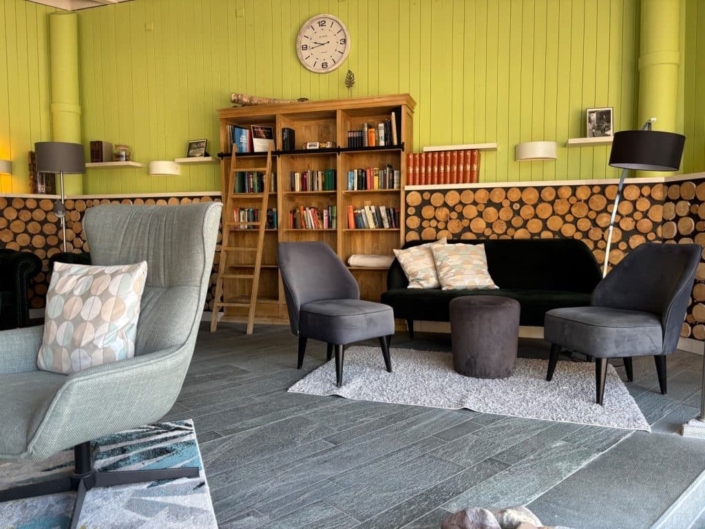 Ein gemütlicher Erholungsraum in der Therme Bad Sassendorf mit hellgrünen Wänden, einem großen Holzregal voller Bücher und verschiedenen Sitzgelegenheiten wie Sesseln und einem Sofa vor einer Wanddekoration aus runden Holzscheiben.