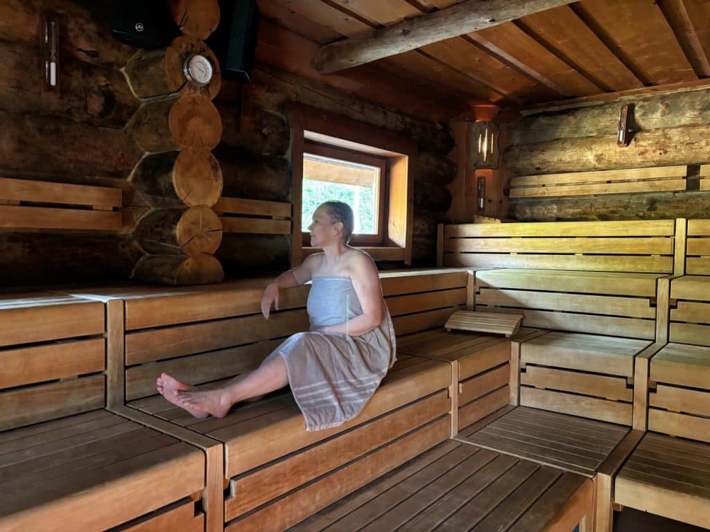 Eine Frau sitzt entspannt in einem Handtuch eingewickelt auf den Holzbänken einer rustikalen Blockbohlensauna in der Börde-Therme Bad Sassendorf und blickt aus einem kleinen Fenster.