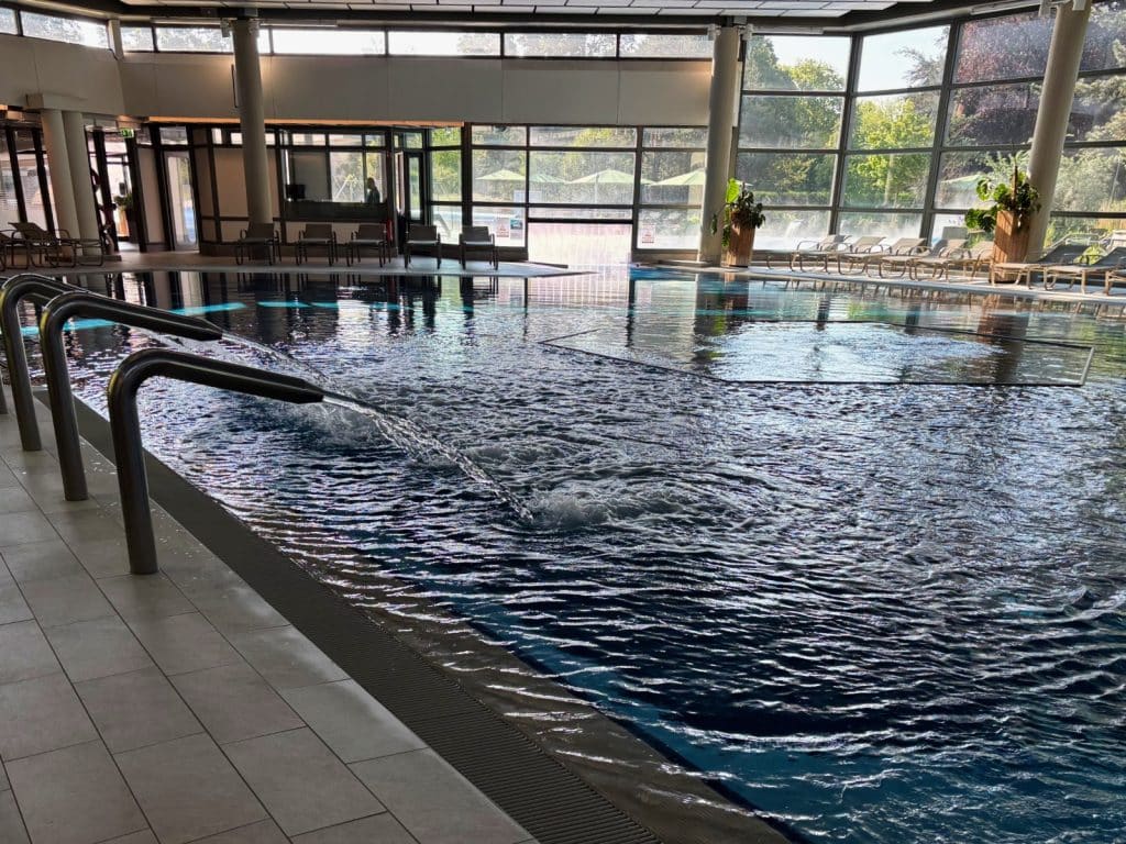 Das Innenbecken der Therme Bad Sassendorf mit sprudelnden Massagedüsen, großen Fensterfronten zum Außenbereich und bereitstehenden Liegestühlen am Beckenrand.