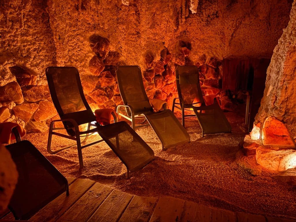 Drei Liegestühle stehen in der rötlich beleuchteten Salzgrotte der Therme in Bad Sassendorf, deren Wände aus groben Salzsteinen bestehen und deren Boden mit feinem Salz bedeckt ist.