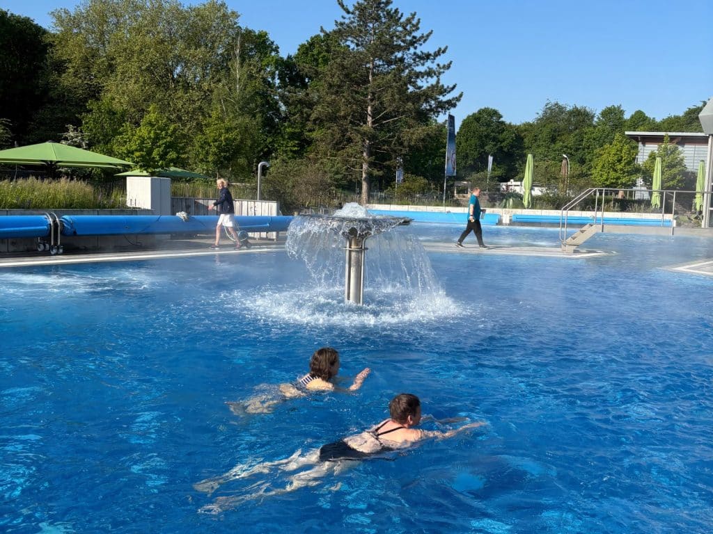 Zwei Frauen schwimmen im beheizten Außenbecken der Börde-Therme in Bad Sassendorf, während im Hintergrund ein Wasserpilz sprudelt und Personen am Beckenrand spazieren.