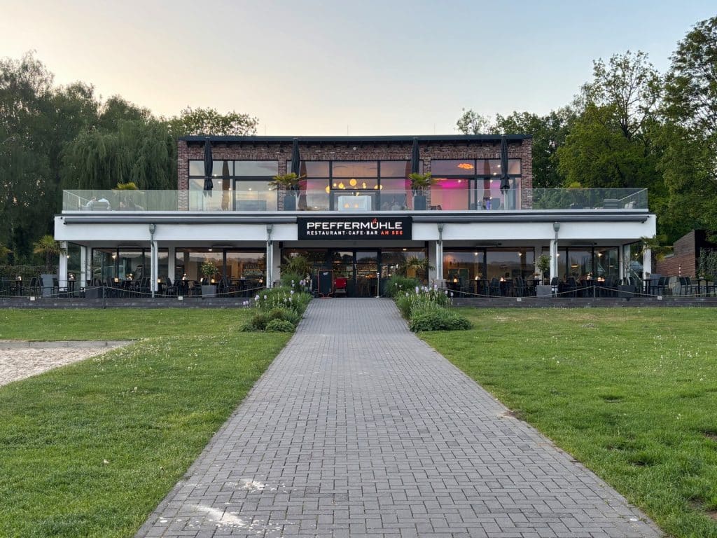 Das zweistöckige Restaurant Pfeffermühle am Möhnesee mit moderner Glasfront und Außenterrasse, zu dem ein gepflasterter Weg durch eine grüne Rasenfläche führt.