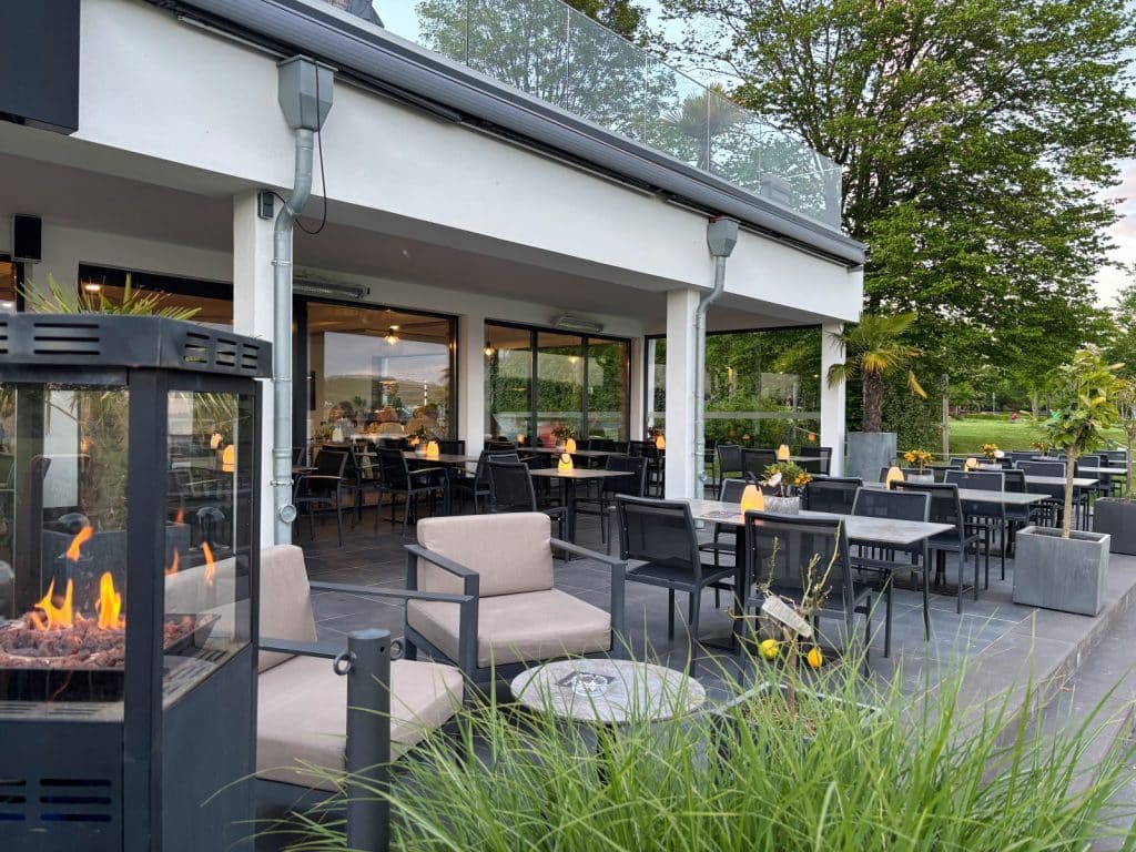 Die Außenterrasse eines Restaurants am Möhnesee mit modernen Lounge-Sesseln im Vordergrund, einem brennenden Heizstrahler und zahlreichen eingedeckten Tischen unter einer überdachten Veranda.