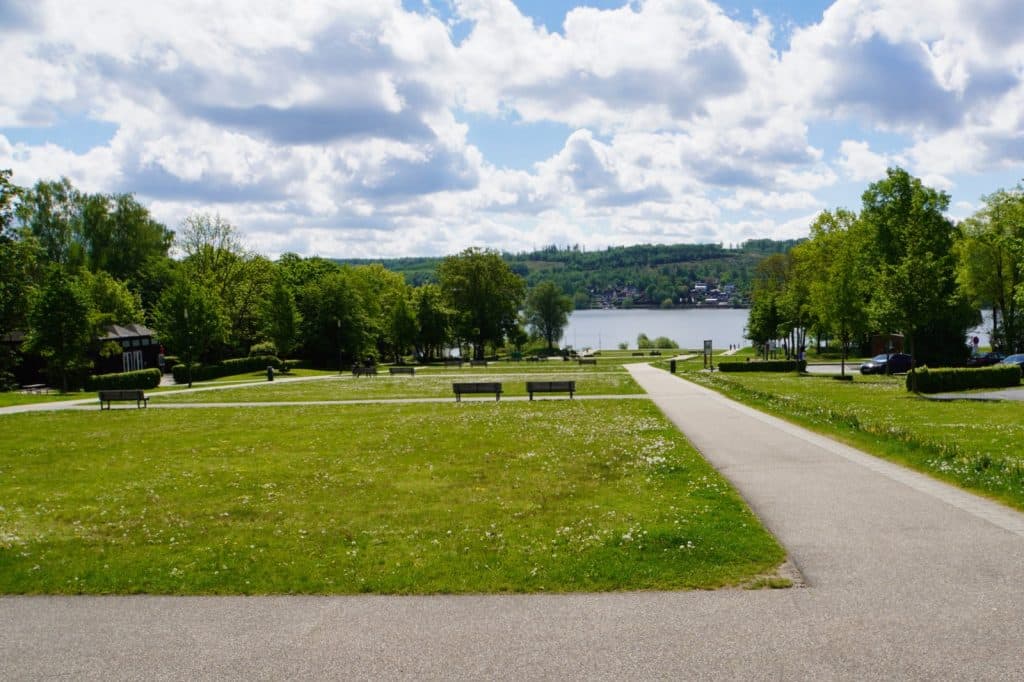 Blick über eine weite Rasenfläche im Seepark Möhnesee mit Parkbänken und gepflasterten Wegen, die auf den glitzernden See unter einem leicht bewölkten Himmel führen.