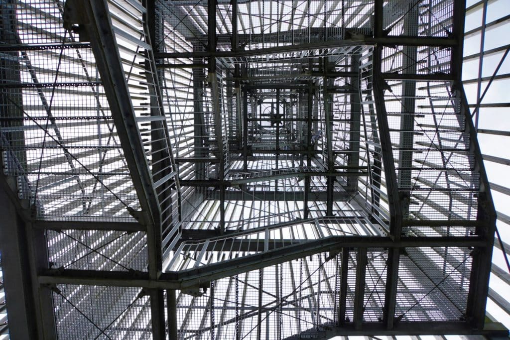 Blick von unten durch die gitterrostartigen Treppenstufen und die Metallkonstruktion des Möhnesee-Turms, die ein komplexes geometrisches Muster aus Linien und Licht bilden.