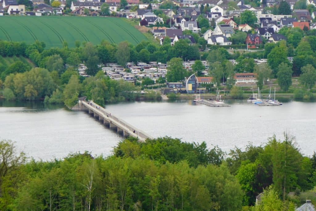 Blick aus der Vogelperspektive auf die Fußgängerbrücke über den Möhnesee, die ein bewaldetes Ufer mit einer Ortschaft, einem Campingplatz und Segelbooten verbindet.