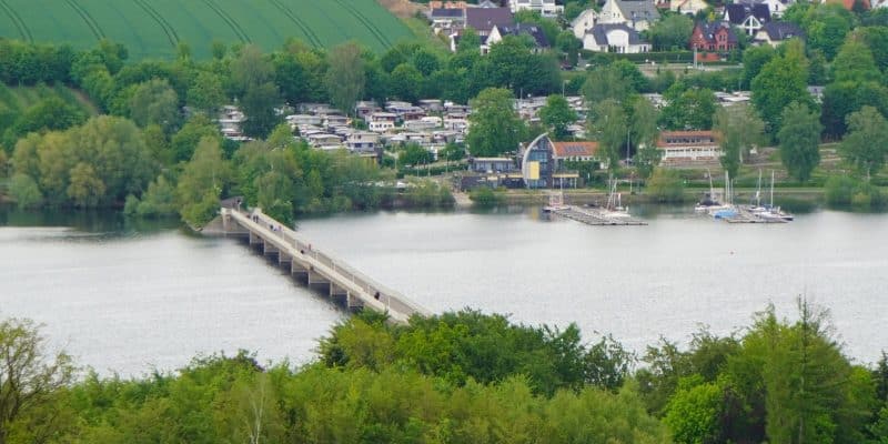 Blick aus der Vogelperspektive auf die Fußgängerbrücke über den Möhnesee, die ein bewaldetes Ufer mit einer Ortschaft, einem Campingplatz und Segelbooten verbindet.