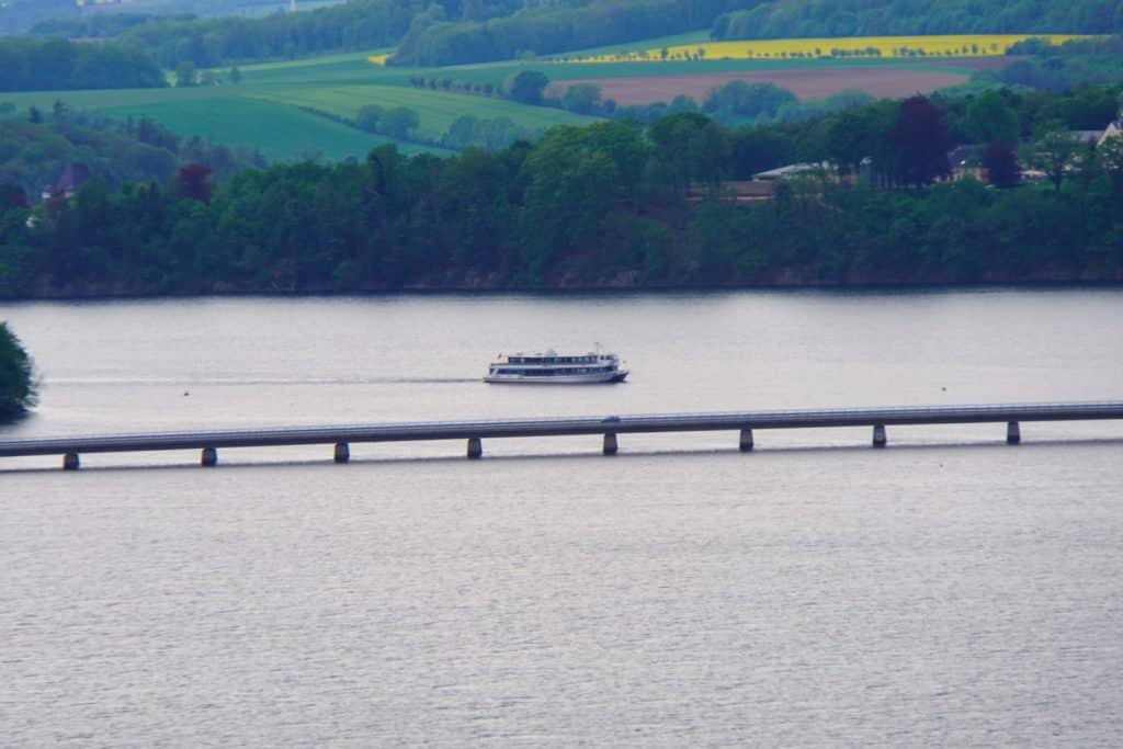 Ein weißes Passagierschiff fährt auf dem Möhnesee hinter einer langen, flachen Brücke, die von zahlreichen Pfeilern gestützt wird, vor einer Kulisse aus bewaldeten Ufern und grünen Feldern.