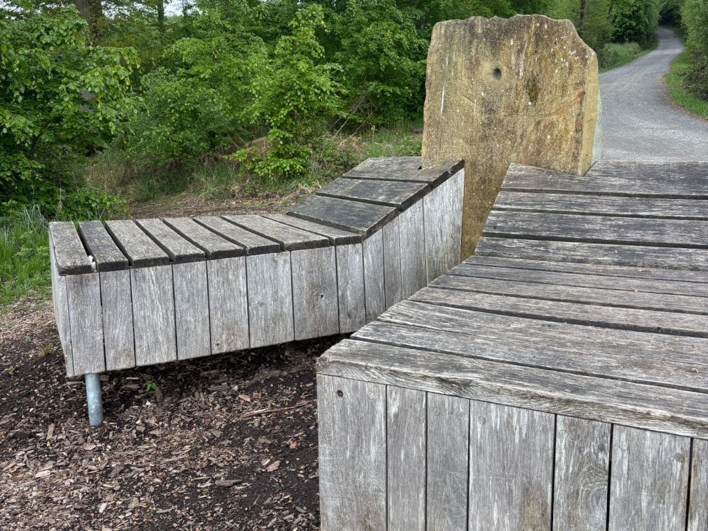 Zwei moderne hölzerne Liegebänke mit einer markanten Rückenlehne aus Naturstein laden am Rand eines Waldweges am Möhnesee zu einer Wanderpause ein.
